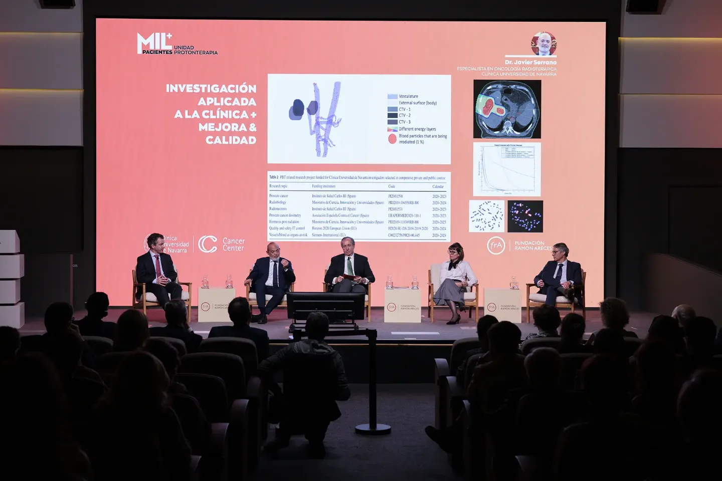 Evento paciente MIL Protonterapia Imagen de los ponentes de una mesa redonda con una proyección en la pantalla que hay detrás de ellos en el evento del paciente MIL de la Unidad de Protonterapia. Clínica Universidad de Navarra