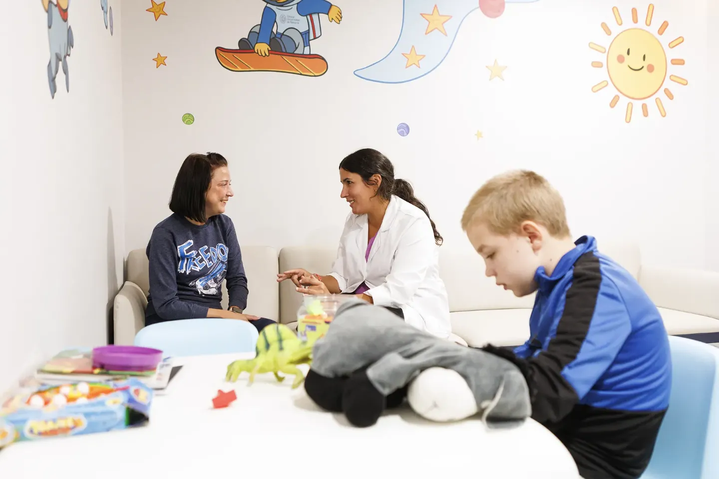 Historia de paciente. Marko y Liu. Beatriz Luquin Imagen de dos mujeres hablando y, en primer plano, un niño sentado a una mesa con juguetes y libros.