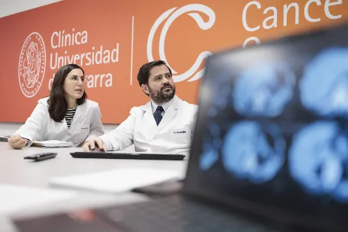 Imagen de un doctor y una doctora con una pantalla de ordenador difuminada en primer plano y el fondo naranja con el logo de la Clínica Universidad de Navarra y el CCUN.