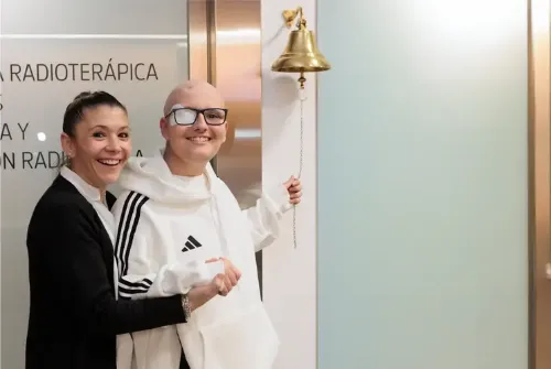 Imagen de Enric, paciente adolescente de la Unidad de Protonterapia de la Clínica Universidad de Navarra, tocando la campana de los valientes acompañado por una auxiliar de enfermería.