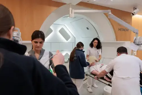 Imagen de Enric, paciente adolescente de la Unidad de Protonterapia de la Clínica Universidad de Navarra, en la sala del gantry junto a auxiliares, técnicos y enfermeras mientras le preparan para el tratamiento.