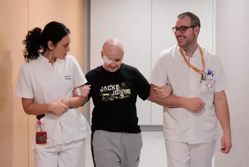 Imagen de Enric, paciente adolescente de la Unidad de Protonterapia de la Clínica Universidad de Navarra, caminando agarrado del brazo dos técnicos de radioterapia por un pasillo del hospital.