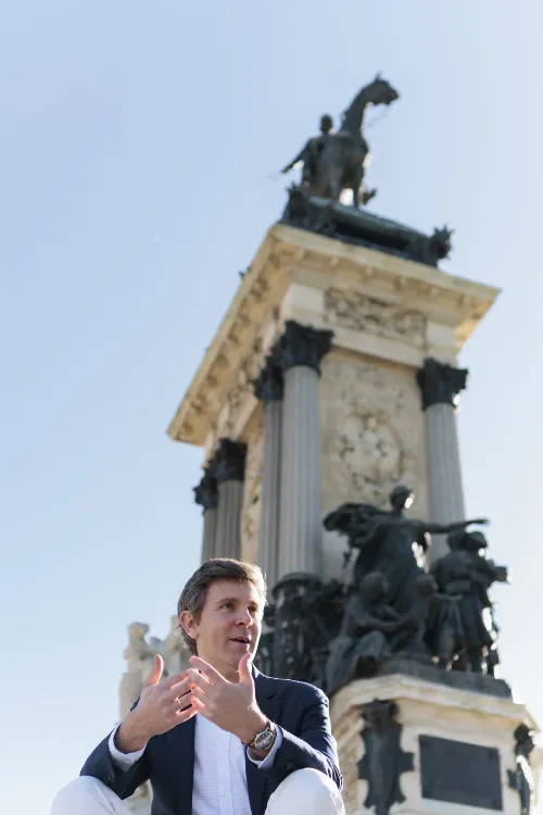Imagen de un médico cardiólogo sin bata sentado en el parque de El Retiro con una gran estatua de fondo.